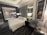 A modern hotel room features a comfortable, neatly arranged bed with white linens. Two contemporary armchairs and a small circular table occupy the corner. A large framed picture of a horse adds character to the room. There is a glass wall with a view into a bathroom, which includes a shower. The room is softly lit by several lamps, and the overall design is sleek and sophisticated.