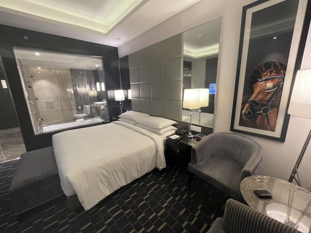 A modern hotel room features a comfortable, neatly arranged bed with white linens. Two contemporary armchairs and a small circular table occupy the corner. A large framed picture of a horse adds character to the room. There is a glass wall with a view into a bathroom, which includes a shower. The room is softly lit by several lamps, and the overall design is sleek and sophisticated.