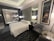 A modern hotel room features a comfortable, neatly arranged bed with white linens. Two contemporary armchairs and a small circular table occupy the corner. A large framed picture of a horse adds character to the room. There is a glass wall with a view into a bathroom, which includes a shower. The room is softly lit by several lamps, and the overall design is sleek and sophisticated.