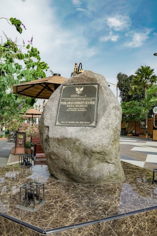 A large rock engraved with a plaque stands prominently in an outdoor setting. The plaque reads 'Pahlawan Street Center Kota Madiun'. Surrounding the rock are benches, greenery including a tree and some potted plants, and a pavilion with a brown roof. The ground is paved with a polished marble-like surface, and a clear blue sky can be seen in the background.