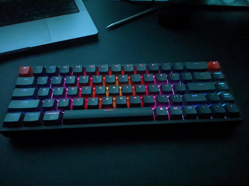 rgb keyboard in a dark room with macbook and metal pen in the background