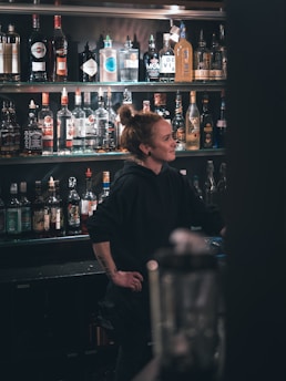 A bartender with a bun hairstyle stands behind a bar, surrounded by a large selection of liquor bottles arranged on shelves. The lighting is dim, creating a cozy atmosphere.