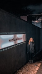 A person stands against a dark wall featuring a glowing sign with the text 'Herzlich willkommen'. The person is wearing a dark jacket with a hood and appears to be casually posed. The scene is dimly lit, with the light focusing on both the person and the sign. The ground is covered with small pebbles.