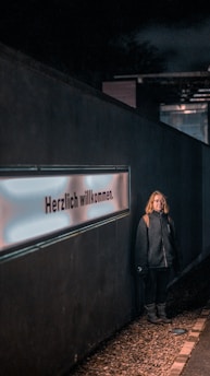 A person stands against a dark wall featuring a glowing sign with the text 'Herzlich willkommen'. The person is wearing a dark jacket with a hood and appears to be casually posed. The scene is dimly lit, with the light focusing on both the person and the sign. The ground is covered with small pebbles.
