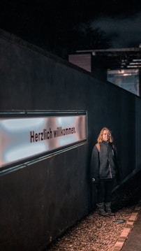 A person stands against a dark wall featuring a glowing sign with the text 'Herzlich willkommen'. The person is wearing a dark jacket with a hood and appears to be casually posed. The scene is dimly lit, with the light focusing on both the person and the sign. The ground is covered with small pebbles.