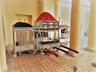 A richly decorated palanquin with an ornate red and black canopy, adorned with intricate designs, is positioned in a historical setting with large columns and a patterned tile floor. Several vintage carriages are visible in the background.