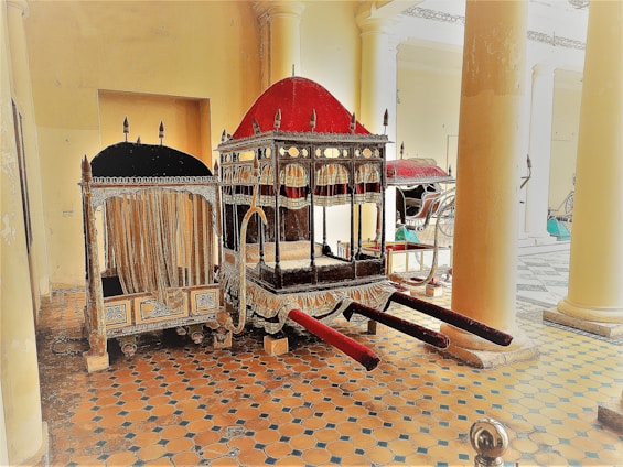 A richly decorated palanquin with an ornate red and black canopy, adorned with intricate designs, is positioned in a historical setting with large columns and a patterned tile floor. Several vintage carriages are visible in the background.
