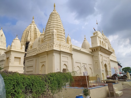 A large, intricately designed cream-colored temple with multiple spires and detailed architecture under a cloudy sky. There is greenery in the foreground and a bundle of wires or ropes near the entrance, with two stone lion statues visible.