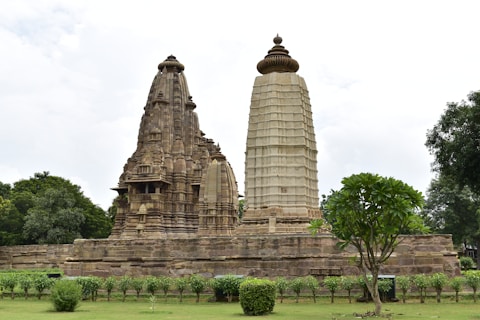 A historic sandstone temple with intricate carvings and a tall, ornate spire surrounded by lush greenery. The temple stands on a raised platform, emphasizing its architectural grandeur, while the sky is overcast, adding a serene atmosphere.
