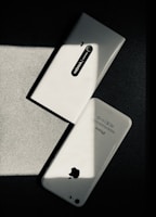 Two white smartphones are laid on a dark surface, intersecting at an angle. One smartphone is a Nokia model with the back camera and model name visible, while the other is an iPhone with the Apple logo and other inscriptions readable. The image has a high contrast with shadow and light creating a striking play.