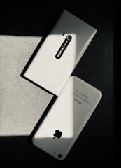 Two white smartphones are laid on a dark surface, intersecting at an angle. One smartphone is a Nokia model with the back camera and model name visible, while the other is an iPhone with the Apple logo and other inscriptions readable. The image has a high contrast with shadow and light creating a striking play.