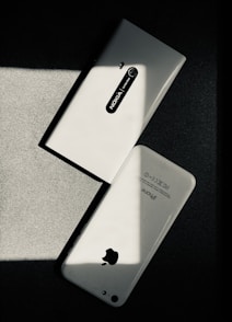 Two white smartphones are laid on a dark surface, intersecting at an angle. One smartphone is a Nokia model with the back camera and model name visible, while the other is an iPhone with the Apple logo and other inscriptions readable. The image has a high contrast with shadow and light creating a striking play.