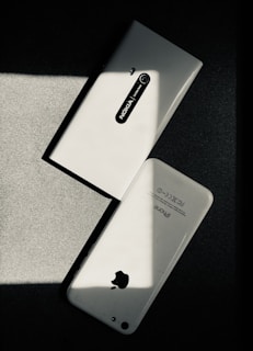 Two white smartphones are laid on a dark surface, intersecting at an angle. One smartphone is a Nokia model with the back camera and model name visible, while the other is an iPhone with the Apple logo and other inscriptions readable. The image has a high contrast with shadow and light creating a striking play.