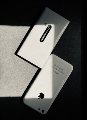 Two white smartphones are laid on a dark surface, intersecting at an angle. One smartphone is a Nokia model with the back camera and model name visible, while the other is an iPhone with the Apple logo and other inscriptions readable. The image has a high contrast with shadow and light creating a striking play.