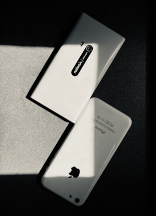 Two white smartphones are laid on a dark surface, intersecting at an angle. One smartphone is a Nokia model with the back camera and model name visible, while the other is an iPhone with the Apple logo and other inscriptions readable. The image has a high contrast with shadow and light creating a striking play.