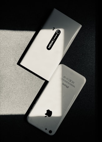 Two white smartphones are laid on a dark surface, intersecting at an angle. One smartphone is a Nokia model with the back camera and model name visible, while the other is an iPhone with the Apple logo and other inscriptions readable. The image has a high contrast with shadow and light creating a striking play.