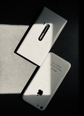 Two white smartphones are laid on a dark surface, intersecting at an angle. One smartphone is a Nokia model with the back camera and model name visible, while the other is an iPhone with the Apple logo and other inscriptions readable. The image has a high contrast with shadow and light creating a striking play.