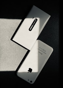 Two white smartphones are laid on a dark surface, intersecting at an angle. One smartphone is a Nokia model with the back camera and model name visible, while the other is an iPhone with the Apple logo and other inscriptions readable. The image has a high contrast with shadow and light creating a striking play.