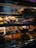 A display case filled with a variety of pastries and baked goods. The top shelf has cakes and pastries, the middle shelf has donuts with various frostings and sprinkles, and the bottom shelf contains croissants and other baked items. Each section is labeled with small signs that indicate the names and prices of the items.