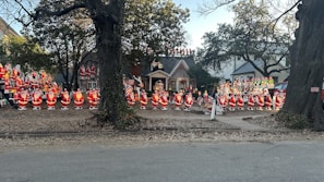 Numerous Santa Claus figures are arranged in a front yard with some additional holiday characters. They are placed in front of a house surrounded by trees, enhancing the festive holiday atmosphere.