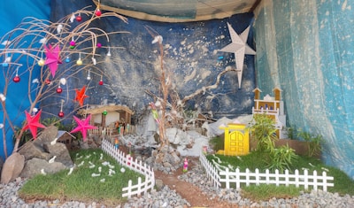 A miniature scene with a nativity setting featuring a small house, various decorations including colorful stars and baubles, green grass patches, white fences, and miniature trees. A blue backdrop with white speckles resembles a starry sky. Rocks and patches of soil add to the natural feel of the scene.