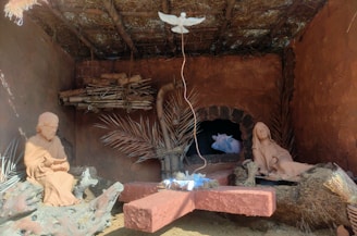 A nativity scene displayed inside a rustic, thatched-roof structure. The scene includes clay sculptures of two figures, one seated and one reclining, with wooden beams and dry palm leaves surrounding them. A cow is visible in an alcove in the background. Above them, a white dove figure is suspended.