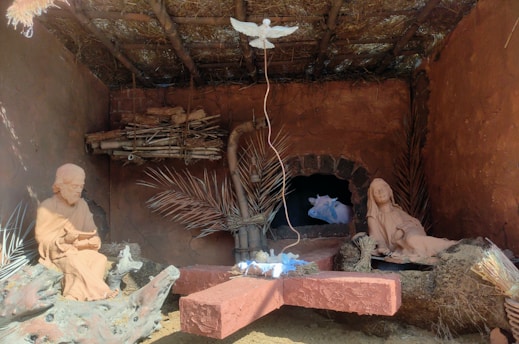 A nativity scene displayed inside a rustic, thatched-roof structure. The scene includes clay sculptures of two figures, one seated and one reclining, with wooden beams and dry palm leaves surrounding them. A cow is visible in an alcove in the background. Above them, a white dove figure is suspended.