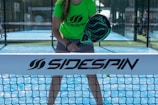 A person stands on a padel court, holding a black padel racket with their body slightly bent forward. They are wearing a bright green shirt with the word 'Sidespin' printed on it, along with gray shorts. The blue court surface is visible, and there is a net in the foreground with the 'Sidespin' logo.