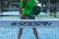 A person stands on a padel court, holding a black padel racket with their body slightly bent forward. They are wearing a bright green shirt with the word 'Sidespin' printed on it, along with gray shorts. The blue court surface is visible, and there is a net in the foreground with the 'Sidespin' logo.