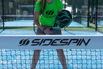 A person stands on a padel court, holding a black padel racket with their body slightly bent forward. They are wearing a bright green shirt with the word 'Sidespin' printed on it, along with gray shorts. The blue court surface is visible, and there is a net in the foreground with the 'Sidespin' logo.
