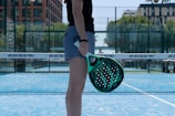 A person is standing on a blue court holding a green and black paddle. There are nets and a backdrop of trees and buildings in the background. The person is wearing a black shirt and grey shorts.