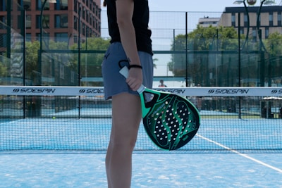 A person is standing on a blue court holding a green and black paddle. There are nets and a backdrop of trees and buildings in the background. The person is wearing a black shirt and grey shorts.