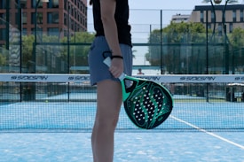 A person is standing on a blue court holding a green and black paddle. There are nets and a backdrop of trees and buildings in the background. The person is wearing a black shirt and grey shorts.