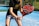 A person is holding a padel racket on an outdoor court with a blue, synthetic playing surface. The racket has a bold design with black, red, and white colors. The individual is dressed in a black shirt with gold lettering and a light gray skirt, suggesting a sporty and focused pose ready for play.