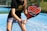 A person is holding a padel racket on an outdoor court with a blue, synthetic playing surface. The racket has a bold design with black, red, and white colors. The individual is dressed in a black shirt with gold lettering and a light gray skirt, suggesting a sporty and focused pose ready for play.
