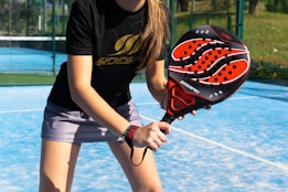 A person is holding a padel racket on an outdoor court with a blue, synthetic playing surface. The racket has a bold design with black, red, and white colors. The individual is dressed in a black shirt with gold lettering and a light gray skirt, suggesting a sporty and focused pose ready for play.