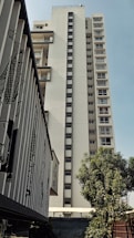 A tall, modern apartment building with multiple floors and balconies. The building has a clean, white facade with vertical lines of windows. In the foreground, there is a smaller structure with decorative panels, and a tree providing some greenery.