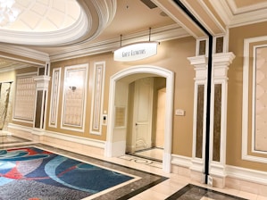 A luxurious hallway with elegant architectural details such as ornate moldings and wall panels. The area is well-lit, with a sign above an arched doorway labeled 'Guest Elevators'. A colorful carpet with abstract patterns lies on the polished floor.