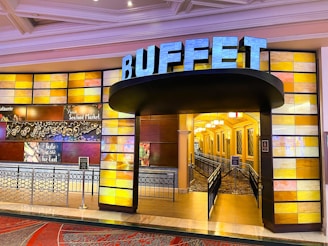 A vibrant entrance to a buffet restaurant featuring a bold sign with the word 'BUFFET' illuminated. The walls are decorated with a mosaic of colorful panels, predominantly in shades of yellow and orange. The interior is visible through the open entrance, showing a warmly lit hallway with decorative light fixtures. Signs on the wall highlight various food options such as rotisserie and seafood market.
