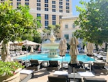 A luxurious outdoor pool area is surrounded by neatly arranged lounge chairs and large parasols. In the center, a decorative fountain sprays water in a symmetrical pattern. Tall, manicured shrubs and flowering plants enhance the setting, with a grand building in the background.