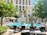 A luxurious outdoor pool area is surrounded by neatly arranged lounge chairs and large parasols. In the center, a decorative fountain sprays water in a symmetrical pattern. Tall, manicured shrubs and flowering plants enhance the setting, with a grand building in the background.