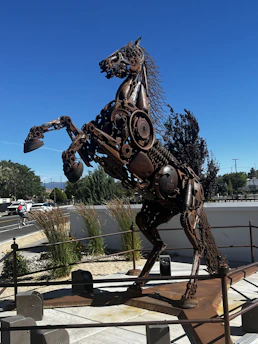 A sculpture of a horse rearing on its hind legs, crafted from various metal parts and mechanical components. The artwork is surrounded by a low fence and positioned in an outdoor setting with clear blue skies. Grasses and shrubs are present in the background, along with a cyclist riding on a nearby road.