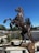 A sculpture of a horse rearing on its hind legs, crafted from various metal parts and mechanical components. The artwork is surrounded by a low fence and positioned in an outdoor setting with clear blue skies. Grasses and shrubs are present in the background, along with a cyclist riding on a nearby road.