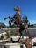 A sculpture of a horse rearing on its hind legs, crafted from various metal parts and mechanical components. The artwork is surrounded by a low fence and positioned in an outdoor setting with clear blue skies. Grasses and shrubs are present in the background, along with a cyclist riding on a nearby road.