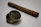 A cigar lies on a flat surface with an open metal tin in the foreground. The tin bears an engraved design with the word 'Amsterdam' prominently displayed. The items are set against a neutral background, enhancing their vintage and luxurious appearance.
