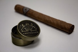 A cigar lies on a flat surface with an open metal tin in the foreground. The tin bears an engraved design with the word 'Amsterdam' prominently displayed. The items are set against a neutral background, enhancing their vintage and luxurious appearance.