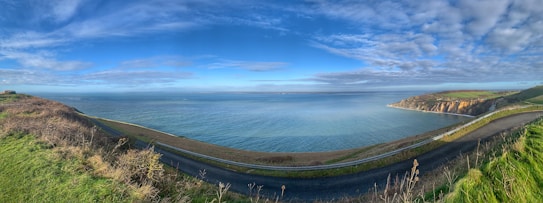 A panoramic coastal landscape featuring a winding road along a cliffside. Verdant green grass covers both sides, with a calm ocean extending to the horizon under a clear blue sky scattered with fluffy clouds. The coastline curves away, revealing rocky cliffs in the distance.