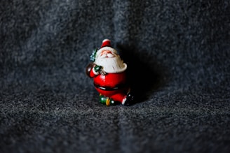 A small figurine of Santa Claus stands against a textured dark gray background. Santa is dressed in his traditional red suit, with white fur trim and black boots. The figurine has a cheerful expression, holding a small green sack over one shoulder.