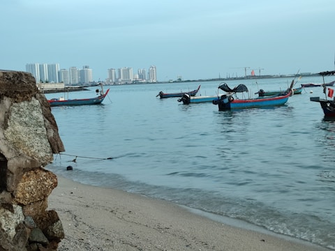 Several small fishing boats are anchored near the shore with a distant view of modern high-rise buildings on the horizon. The water is calm, reflecting the clear blue sky, and the shoreline is composed of fine sand and a rocky foreground.