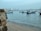 Several small fishing boats are anchored near the shore with a distant view of modern high-rise buildings on the horizon. The water is calm, reflecting the clear blue sky, and the shoreline is composed of fine sand and a rocky foreground.
